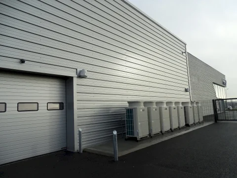 Rear exterior view of the Audi dealership workshop at Futura Park, Ipswich, showcasing Kingspan RW composite wall panels in light grey. The image features a large sectional garage door with built-in vision panels, multiple HVAC units aligned along the wall, LED security lights, and a black security gate leading to the service yard.