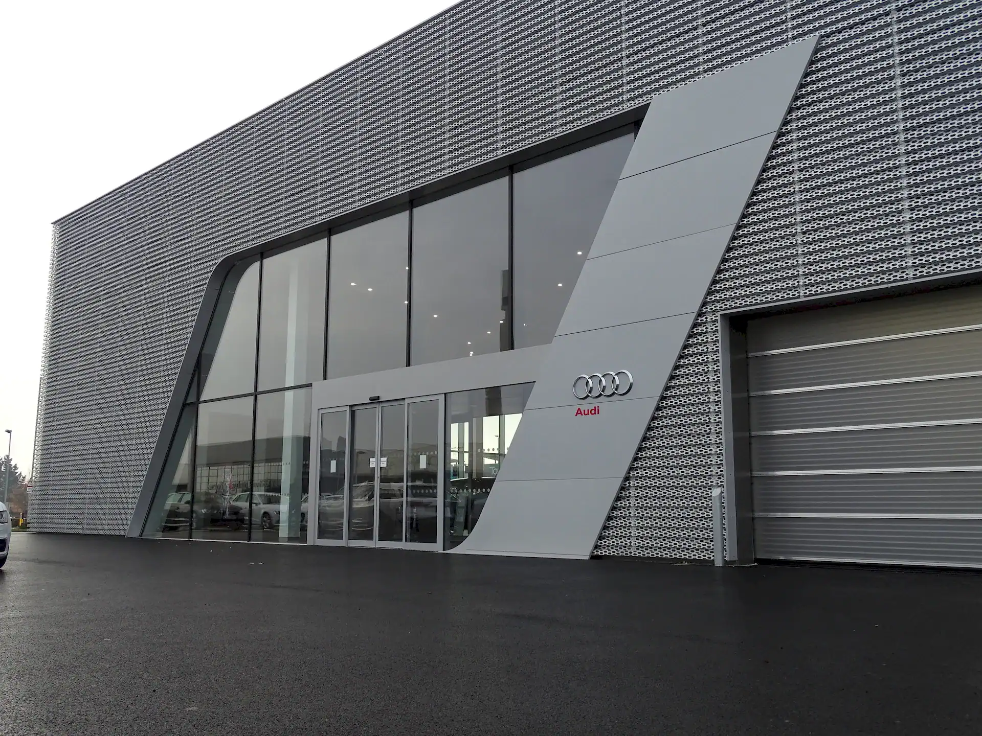 Exterior view of the Audi dealership at Futura Park, Ipswich, Suffolk, showcasing the modern facade. The building features a perforated anodised aluminium rainscreen cladding installed on an aluminium bar and bracket framework. The entrance has large floor-to-ceiling glass windows framed by angled aluminium panels, with the Audi logo displayed prominently. To the right, a large grey roller shutter door provides access to the workshop area. The surrounding area consists of freshly laid black asphalt, reflecting the dealership's premium finish.
