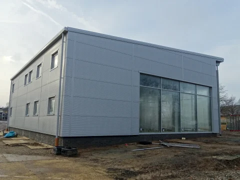 A rear-side view of the Orwell Truck & Van Centre under construction, showing the completed composite cladding system. The building features silver-grey Tata Steel Trimapanel wall panels with a seamless vertical joint design. Large floor-to-ceiling glass windows are visible on the right side, reflecting trees and the surrounding area. The foundation is built with a dark brick base, and construction materials, including pipes and wooden planks, are scattered around the site. The background features a cloudy sky and a partially visible construction area.