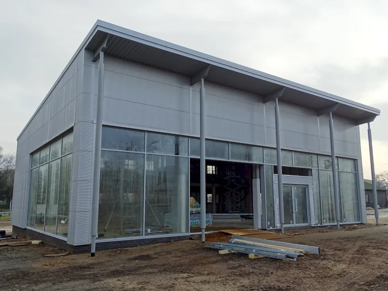 A modern, partially constructed van sales centre with a sleek grey Tata Steel Trimapanel composite wall exterior. The building features a large glass-fronted façade with full-height windows and a double glass entrance door. The extended roof canopy is supported by slim steel columns, with visible guttering along the edge. The construction site in the foreground has an unfinished dirt surface, construction materials, and scattered metal beams. The interior of the building is visible through the glass, showing an unfinished framework. The sky is overcast, and trees line the background.