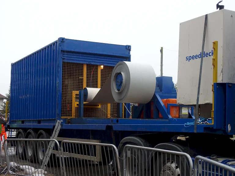 Side view of the SpeedDeck mobile rolling unit in operation at the Two Rivers Medical Centre construction site. A large coil of steel is mounted on a blue trailer, feeding into the SpeedDeck forming machine, which shapes the material into standing seam roof panels. The machine is surrounded by safety barriers, with construction workers monitoring the process in the background.