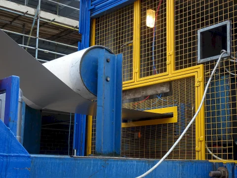 Close-up of the SpeedDeck mobile rolling unit in operation at the Two Rivers Medical Centre construction site. A large coil of steel is mounted on a blue de-coiler stand, feeding into the yellow-framed rolling machine, where it is formed into standing seam panels. The protective mesh guards the internal machinery, and a white cable runs along the side.