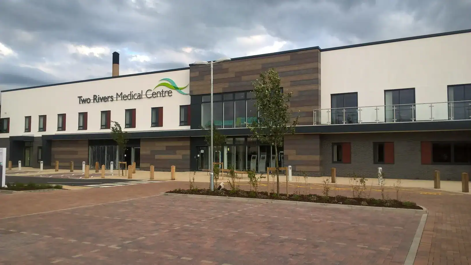 A completed Two Rivers Medical Centre building with a modern facade featuring white and wood-textured rainscreen panels. The entrance has glass double doors, with a balcony on the upper level that has glass railings. The building name and logo are prominently displayed on the upper facade. The surrounding area includes a landscaped walkway, young trees, and brick-paved parking spaces under a cloudy sky.