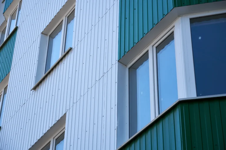 Close-up of a modern building facade featuring Twin-Skin Built-Up Walling with green and white corrugated metal panels and protruding windows. The cladding system enhances both insulation and weather resistance.