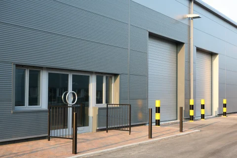 Modern commercial building exterior featuring twin-skin built-up walling installed by S & G Industrial Roofing. The walls consist of horizontal grey steel panels with large dark-blue roller doors, pedestrian doors, and lighting, ideal for offices and commercial premises.